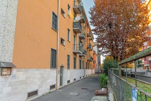 Eine schmale Stadtstraße mit mehrstöckigen Gebäuden, einem baumbewachsenen Bürgersteig und einem Metallzaun.