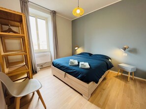 Quarto com cama, mesas de cabeceira, uma cadeira, guarda-roupa e janela.