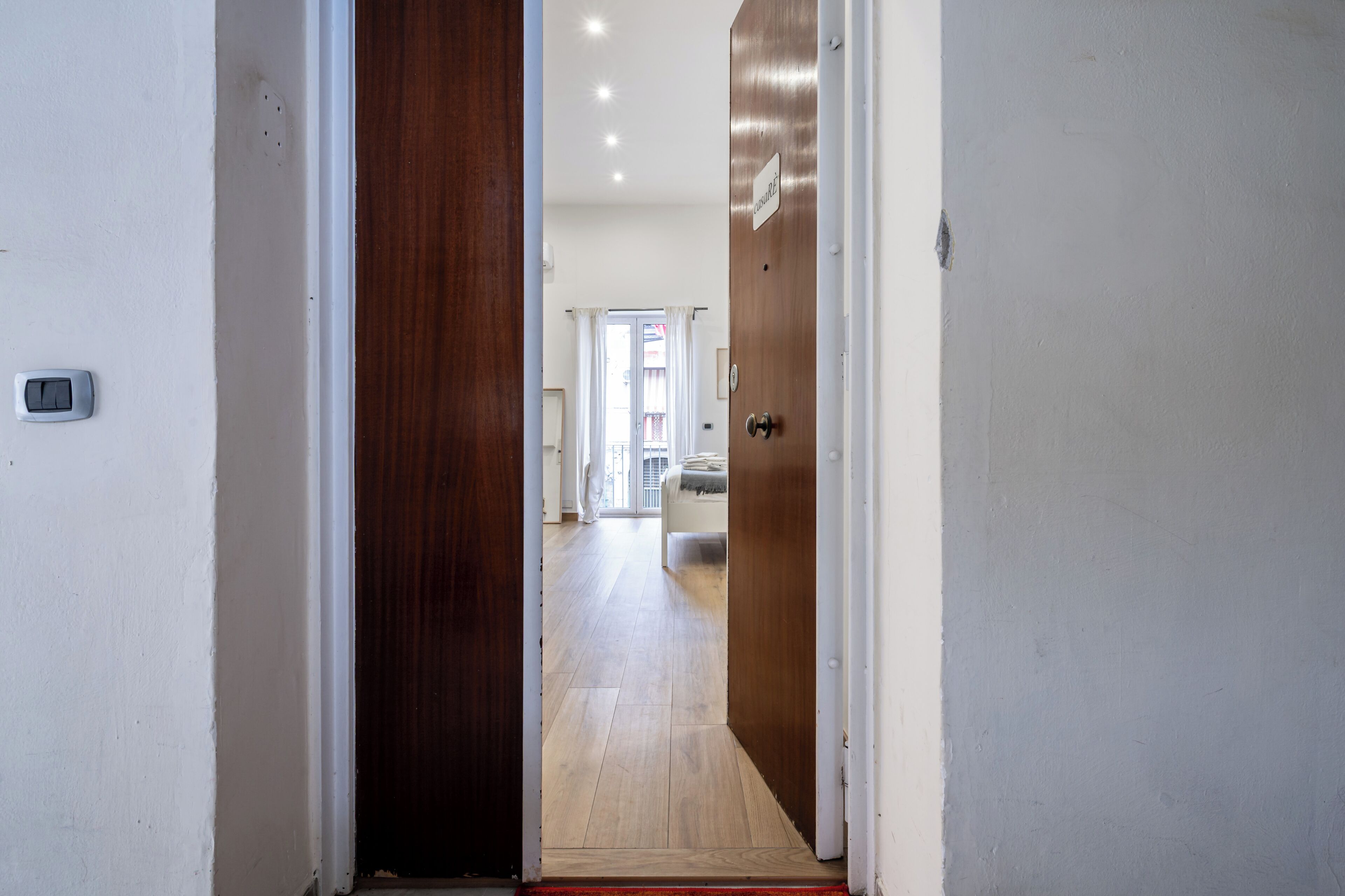 Un pasillo con una puerta de madera, un interruptor de luz y vista a una habitación con cama y ventana.