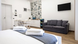 A modern hotel room with a bed, grey sofa, flat-screen TV, and a small kitchenette. - Casaré (Naples)