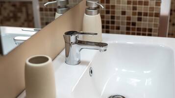 Ein Badezimmerwaschbecken mit chromfarbenem Wasserhahn, einem beigen Seifenspender und einem Spiegel, der eine geflieste Wand reflektiert.