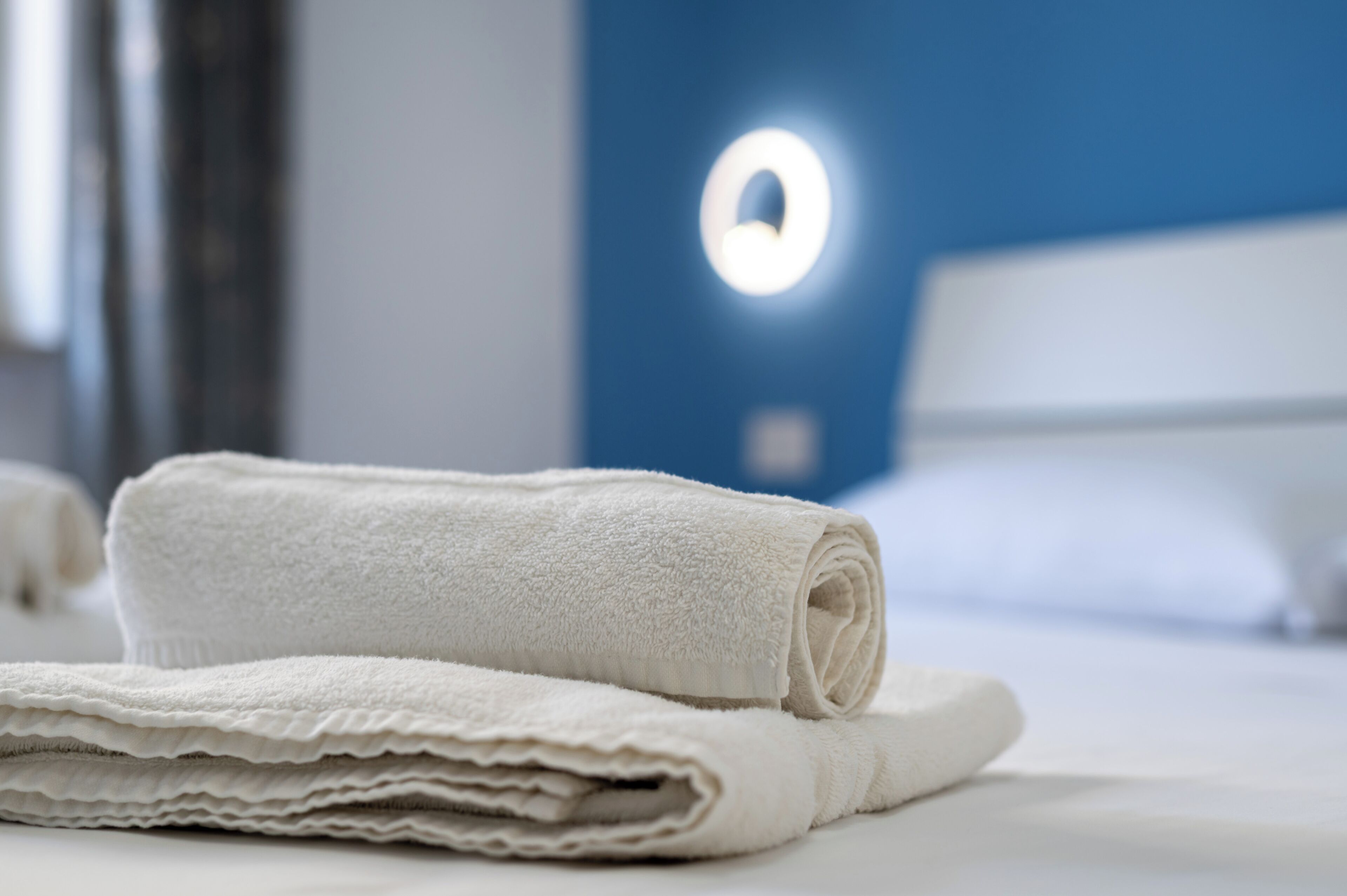 Een keurig opgevouwen witte handdoek op een bed, met een blauwe wand en een ronde lamp in de achtergrond.