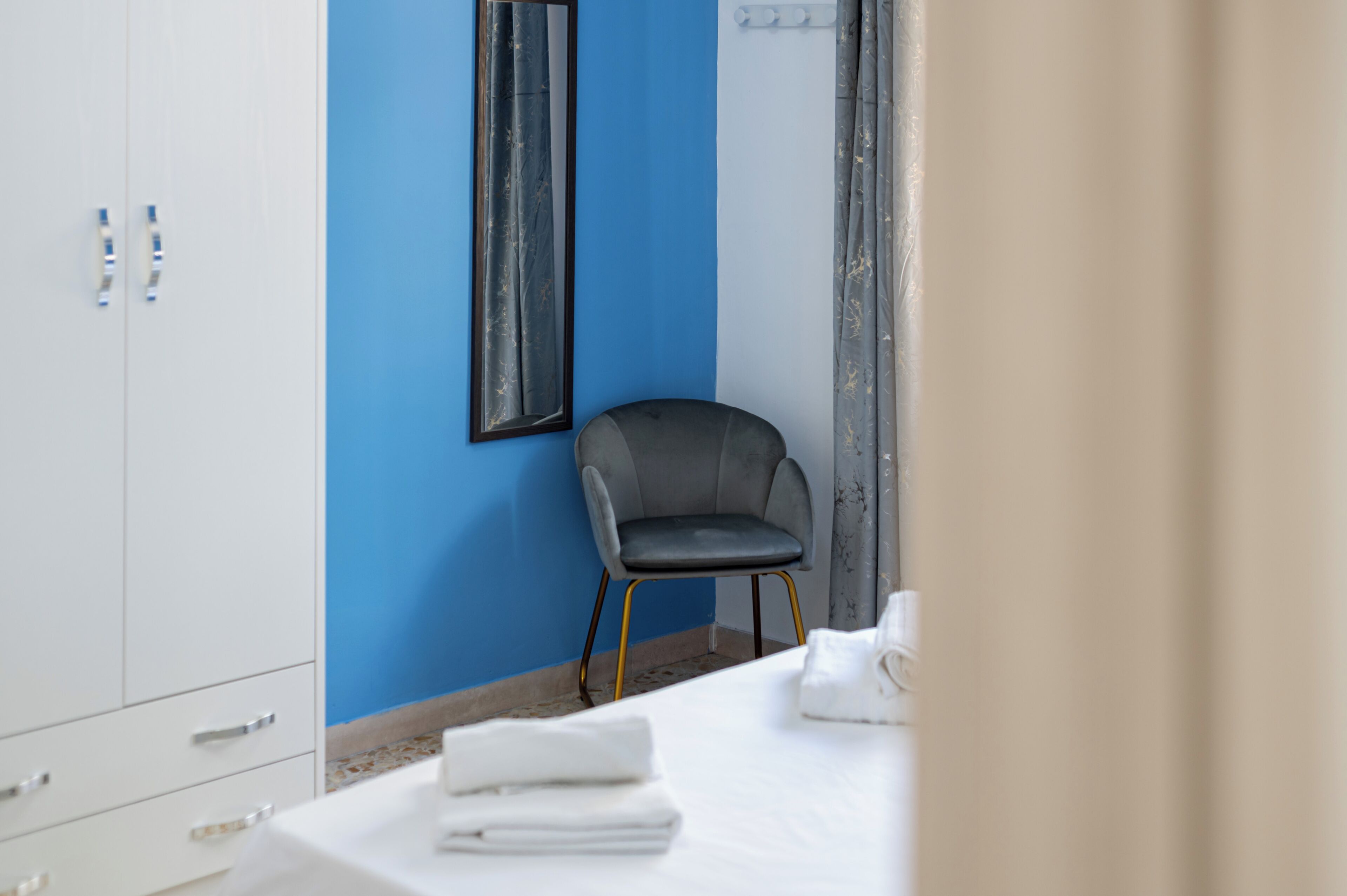 A hotel room with a blue accent wall, a grey upholstered chair, a white table with folded towels, and a large mirror.