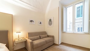 Habitación de hotel con un sofá color beige, una mesita de noche con lámpara y una ventana con vistas a edificios.