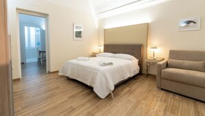 Un dormitorio con cama, mesitas de noche, una silla y una puerta que da a otra habitación.