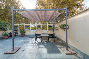 Ein überdachter Patio-Bereich mit Tisch und Stühlen, umgeben von Kübelpflanzen und einer Pergola mit Dach.