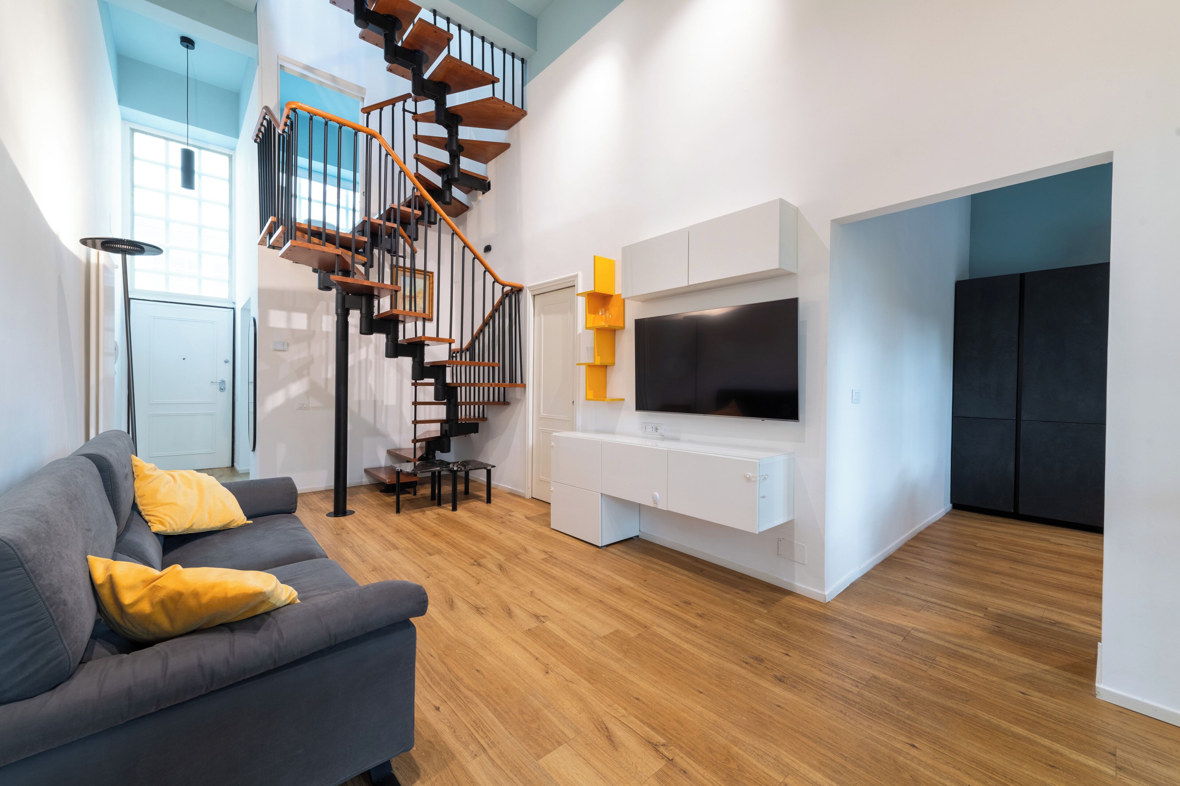 A modern living room with a grey sofa, yellow cushions, a wooden staircase, and a flat-screen TV mounted on a white cabinet.