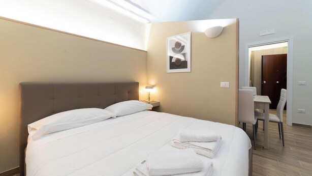 Un lit bien fait, avec du linge de lit blanc et des oreillers, une lampe de chevet, un tableau encadré au mur et un coin repas avec une table et des chaises.