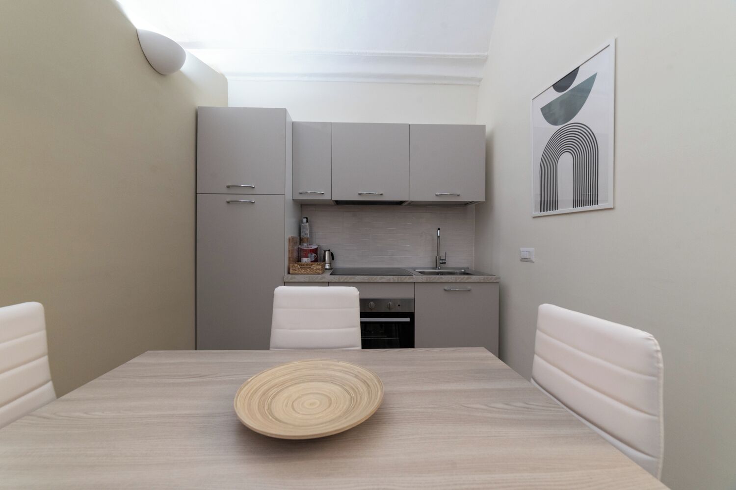 Une cuisine compacte avec des meubles gris, un plan de travail blanc et un évier. On y trouve une table à manger avec un plateau en bois au centre.