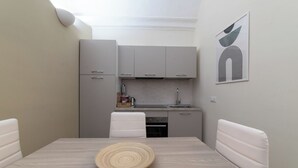 Une cuisine compacte avec des meubles gris, un plan de travail blanc et un évier. On y trouve une table à manger avec un plateau en bois au centre.