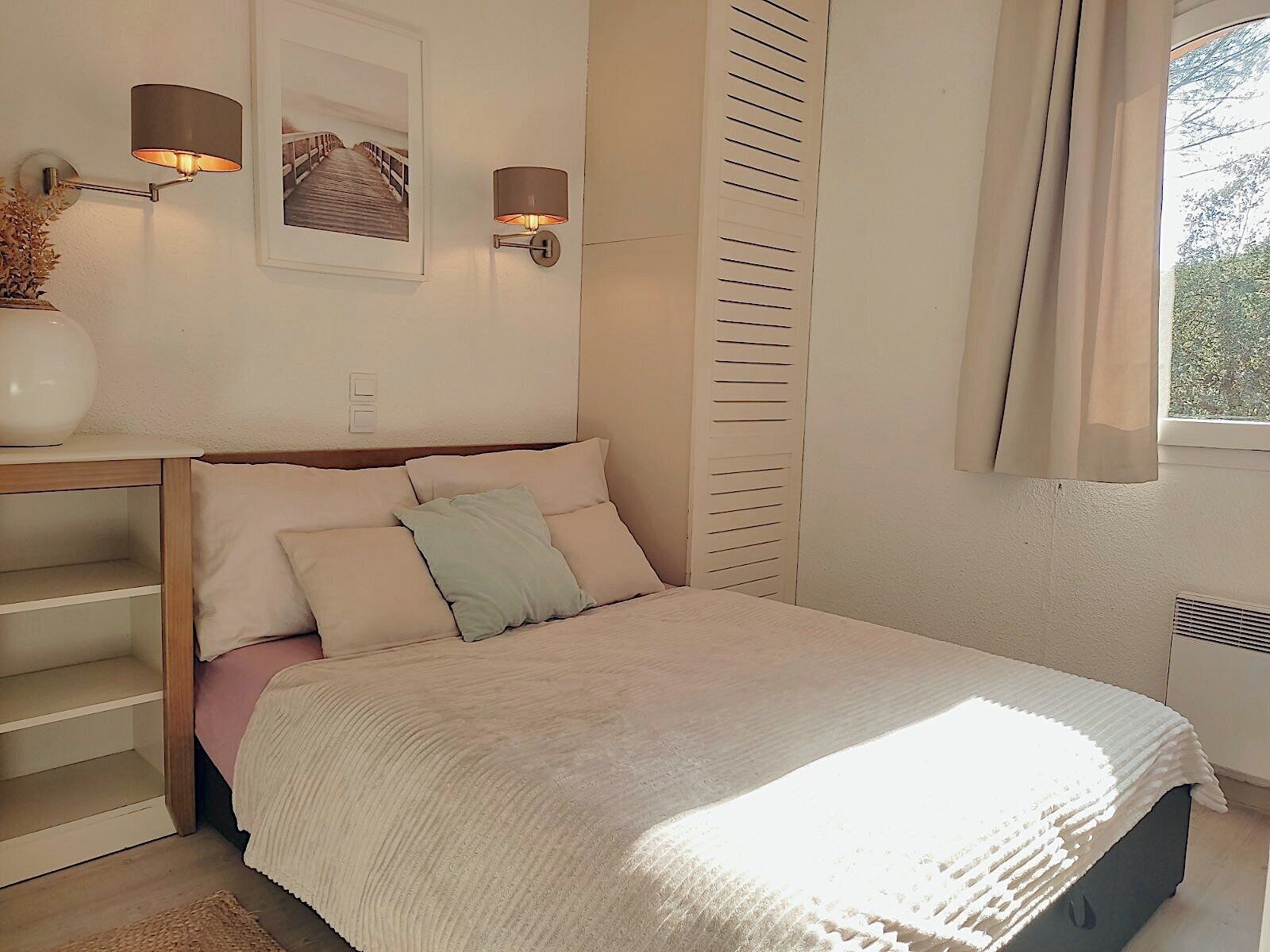 Un dormitorio con una cama, dos lámparas de pared, un cuadro, una ventana con cortinas y un radiador.