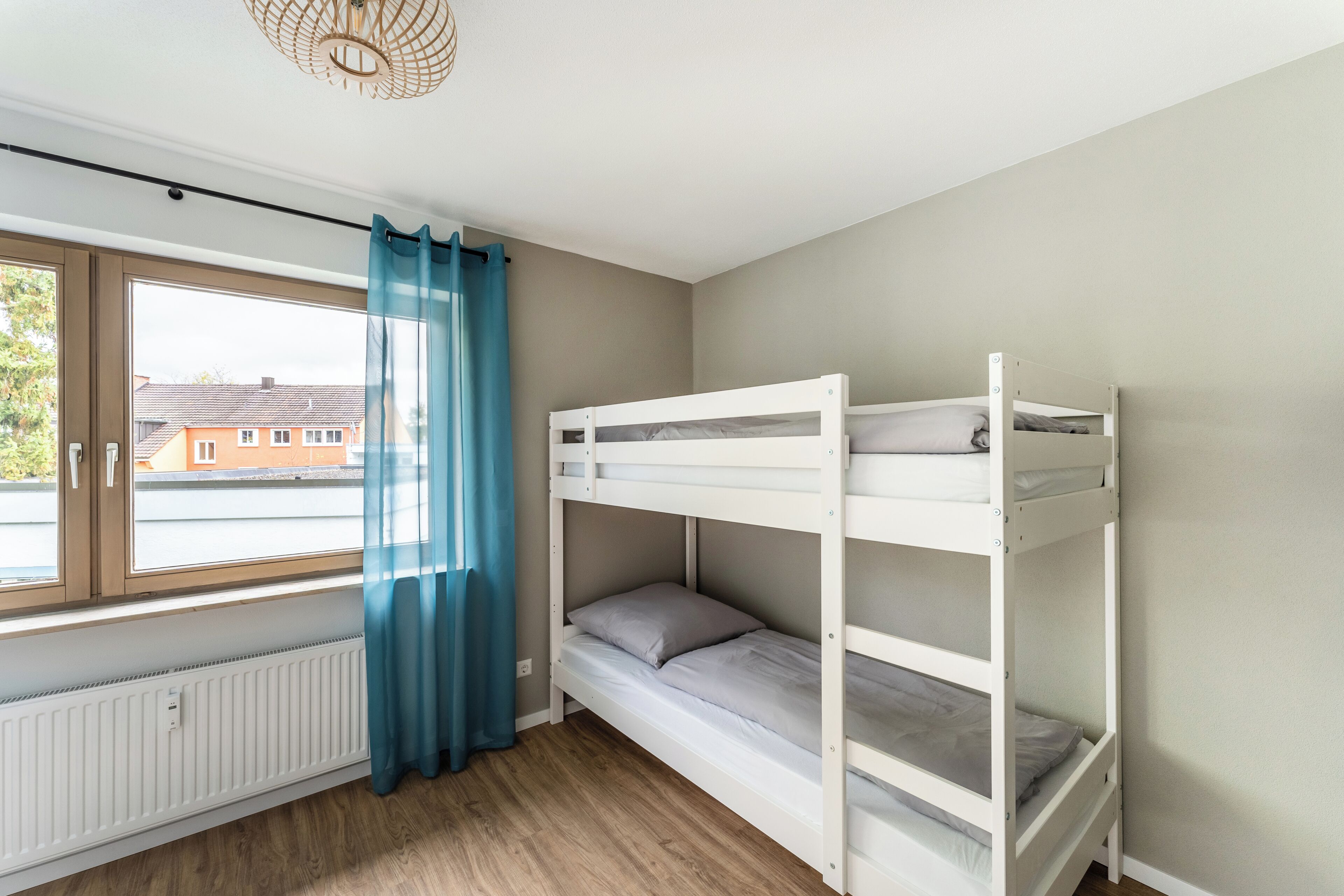 A room with a bunk bed, a window with curtains, a radiator, and a view of houses outside.