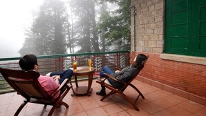 Dos personas sentadas en sillas de madera alrededor de una mesa redonda pequeña, con bebidas, mirando un balcón con una puerta verde y vistas a árboles.