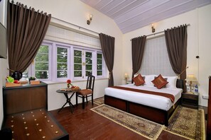 A hotel room with a bed, a desk, a chair, a small table with a vase and fruit, and two windows with curtains. - Brij Villa Dalhousie A Colonial Luxury Retreat (Dalhousie)