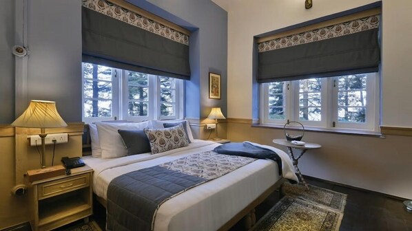 A bedroom with two beds, large windows with dark curtains, bedside lamps, a bedside table, and a small round table with a mirror. - Brij Villa Dalhousie A Colonial Luxury Retreat (Dalhousie)