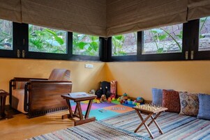 A room with a wooden chair, a small table, a sofa, and a chessboard. There are windows with a view of greenery outside.