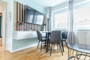 Uma cozinha moderna com fogão embutido, área de refeições com mesa e cadeiras de vidro, e vista para edifícios através de uma grande janela.
