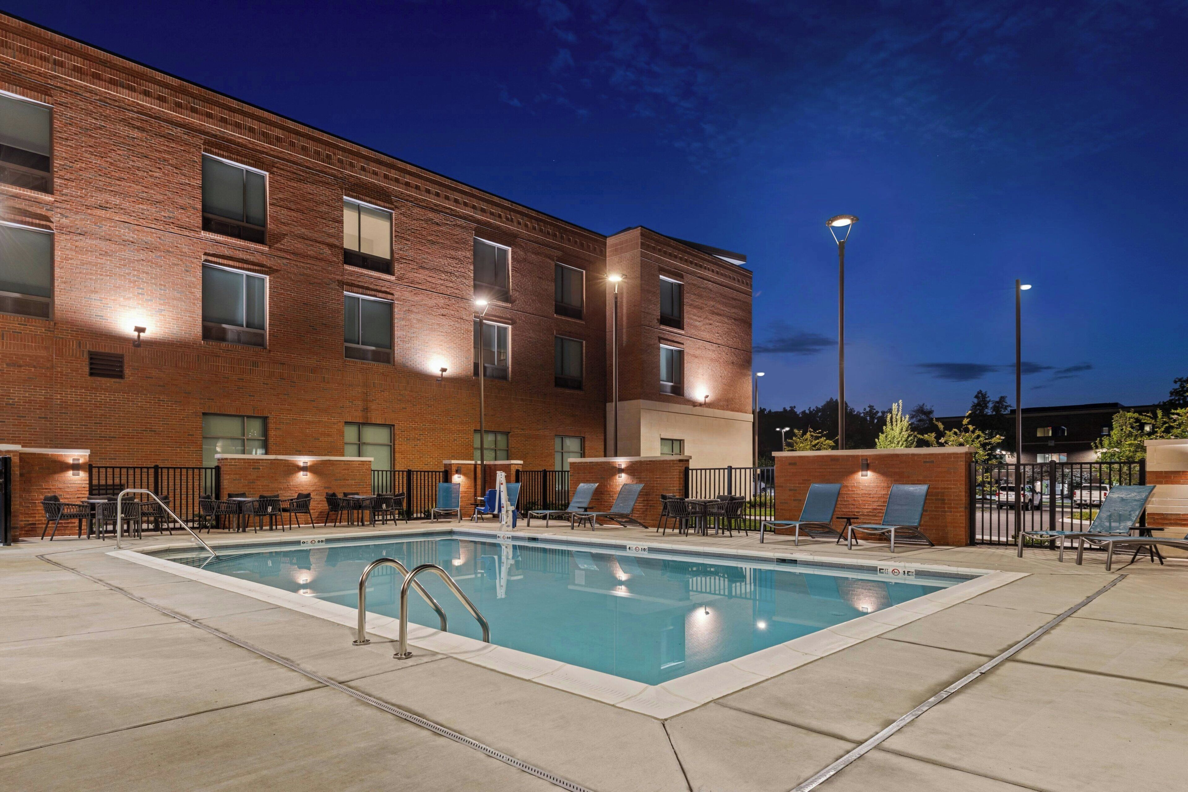 Una piscina con sillas de descanso y mesas, rodeada por un edificio de ladrillo con amplias ventanas e iluminación exterior.