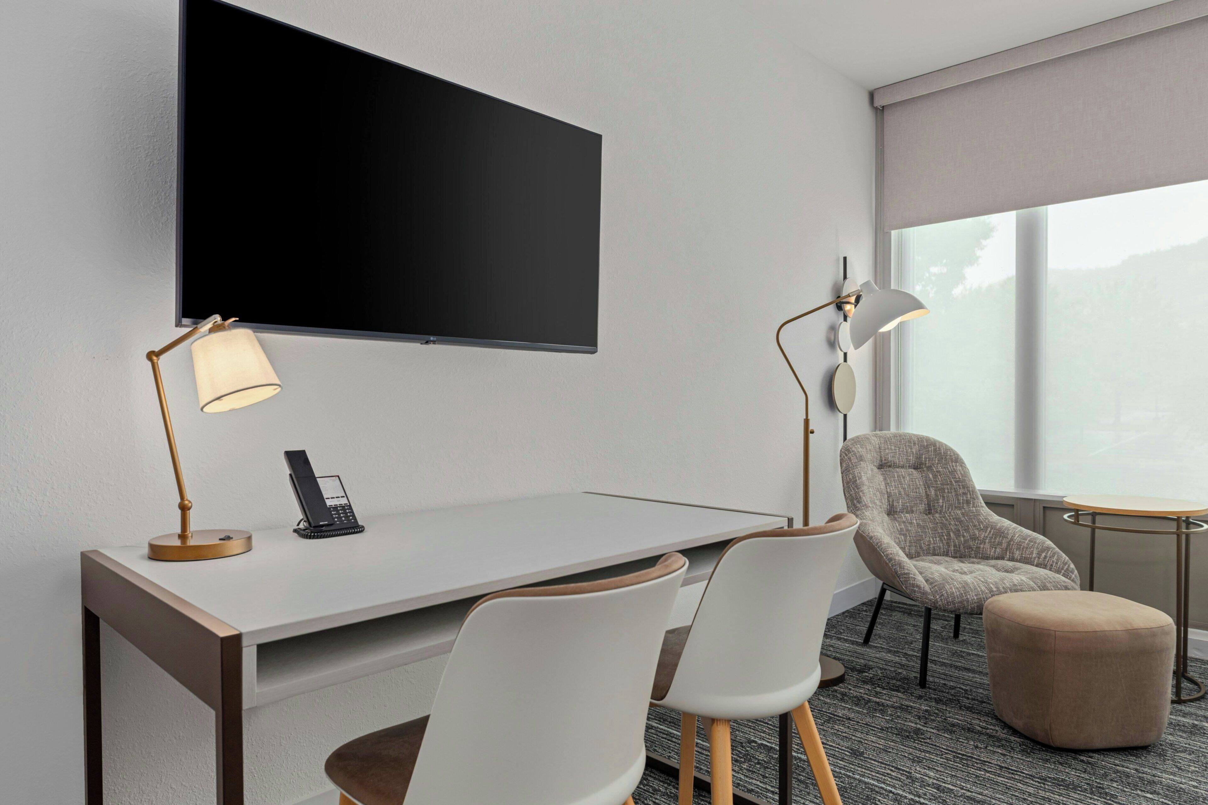 Quarto de hotel com televisão de tela plana na parede, escrivaninha com telefone e luminária, duas cadeiras, um pequeno banquinho e janela com vista.