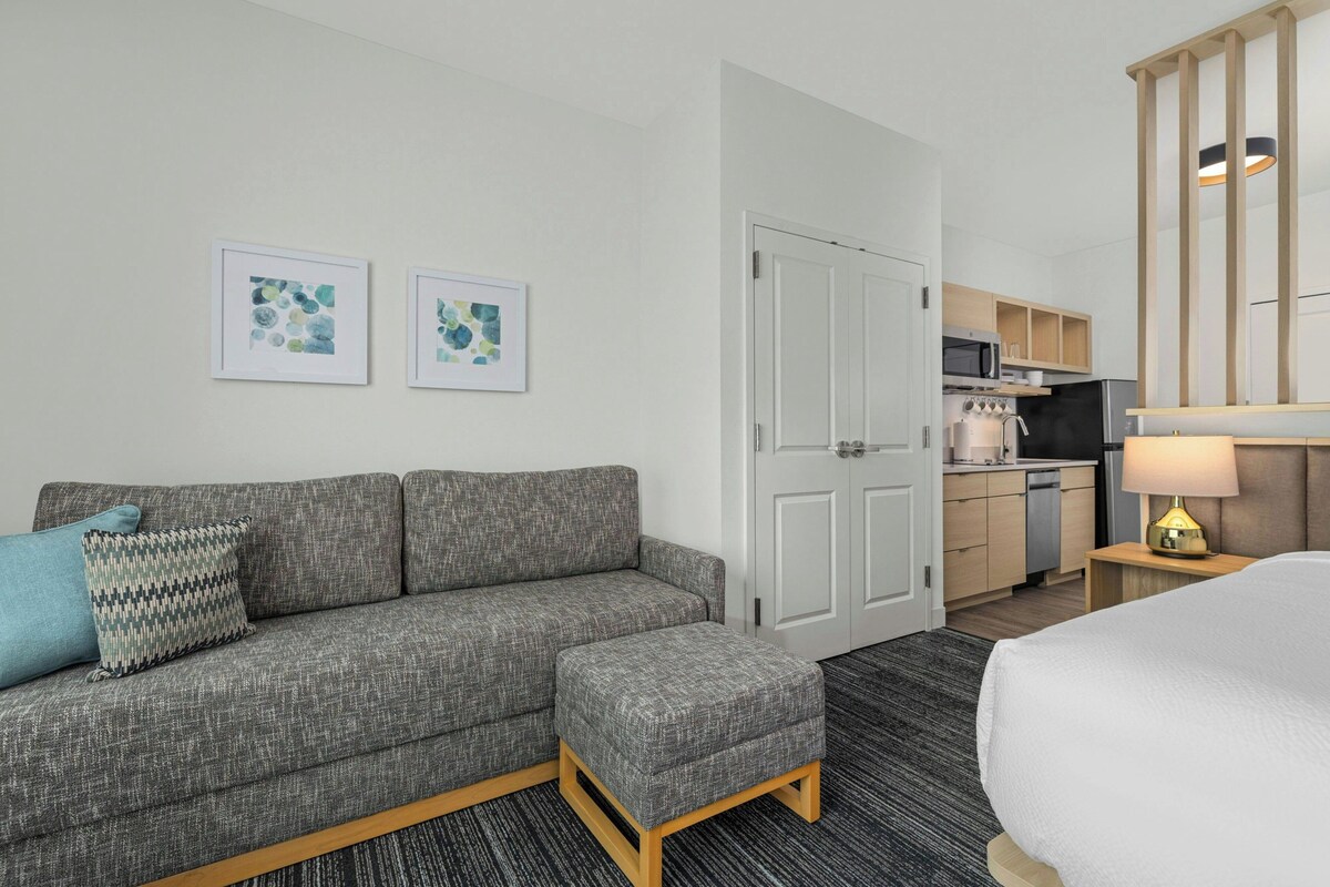 A hotel room with a grey sofa, a matching ottoman, a bed, a kitchenette, and two framed artworks on the wall.