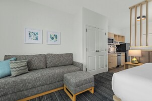 Habitación de hotel con un sofá gris, un reposapiés a juego, una cama, una kitchenette y dos cuadros en la pared.