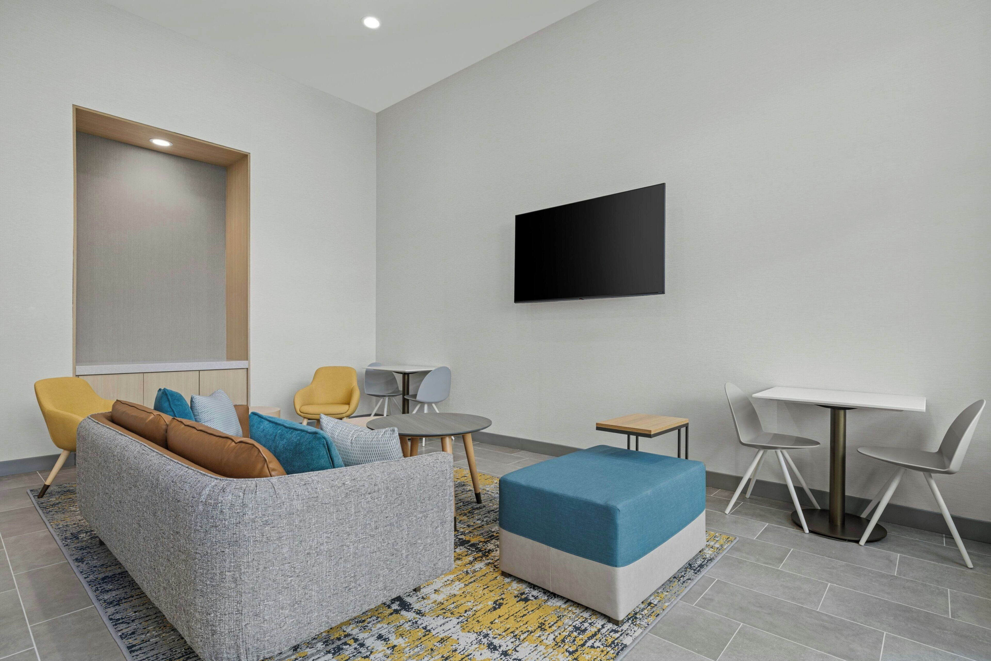 A modern living room with a grey sofa, yellow and blue chairs, a small round table, a blue ottoman, and a flat-screen TV mounted on the wall.