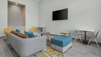 A modern living room with a grey sofa, yellow and blue chairs, a small round table, a blue ottoman, and a flat-screen TV mounted on the wall.