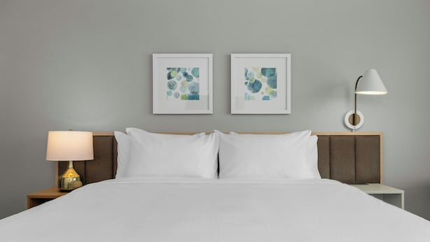 Quarto de hotel com uma cama, duas pinturas abstratas emolduradas, uma luminária de cabeceira e uma mesa de apoio.