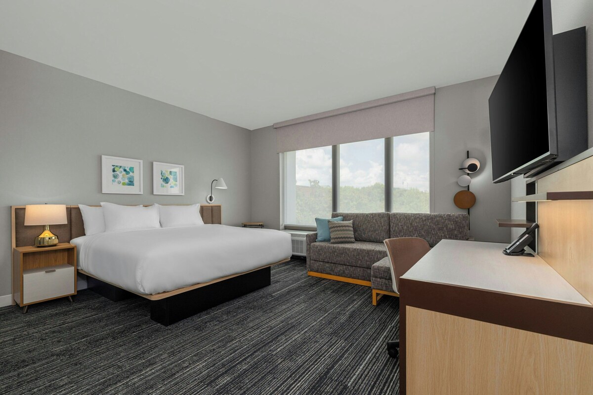 A hotel room with a bed, a desk, a TV, a sofa, and artwork on the wall.