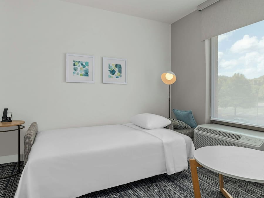 Quarto de hotel com cama, mesa redonda, luminária de pé e duas obras de arte emolduradas na parede.