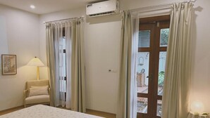 A bedroom with a bed, a chair, an air conditioner, a door leading outside, and a small table with a lamp. - In Home (Hanoi)