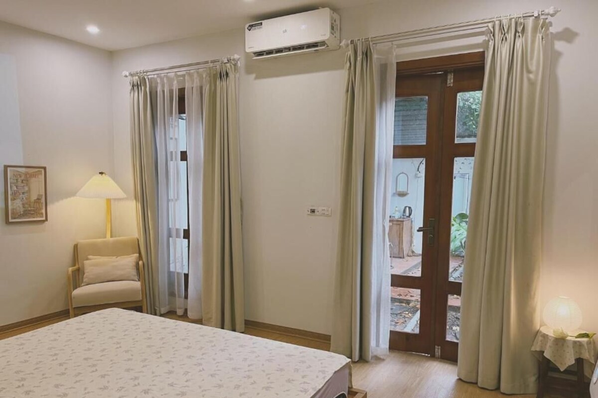 A bedroom with a bed, a chair, an air conditioner, a door leading outside, and a small table with a lamp.