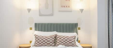 Una cama bien hecha con almohadones estampados, un cabecero tapizado en verde y dos lámparas de noche.