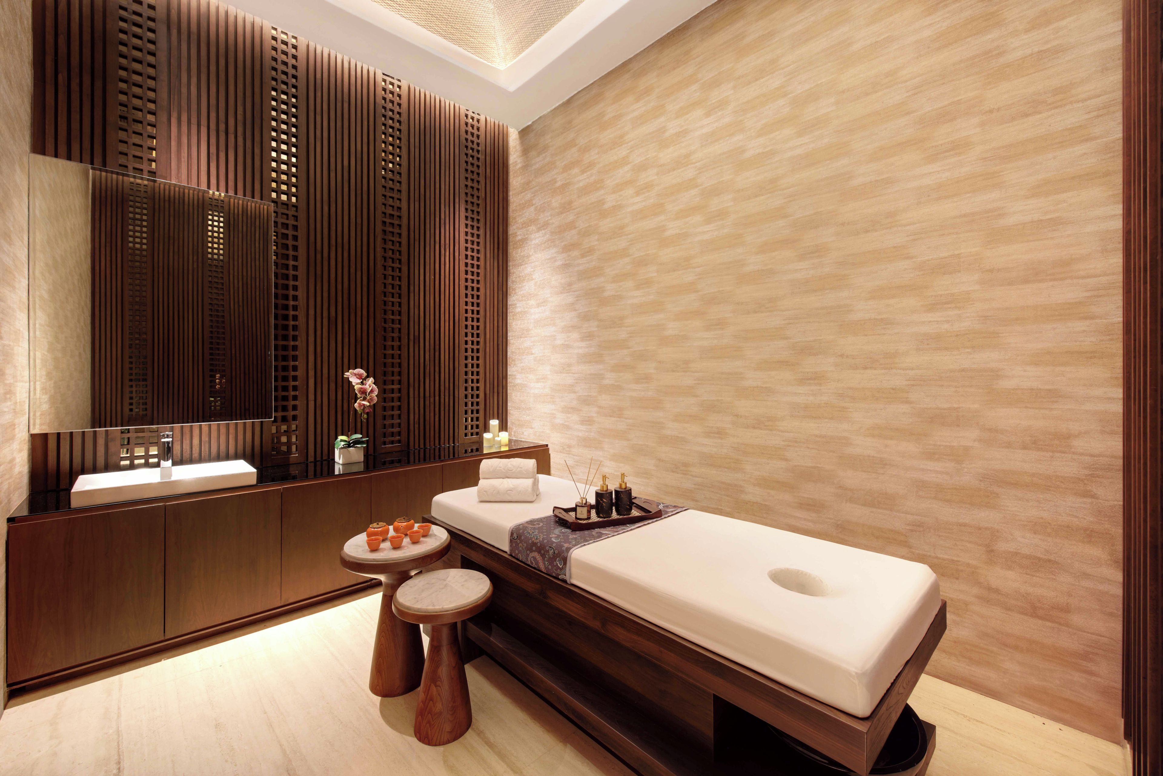 A modern spa room with a wooden panel wall, a large mirror, a massage table, a small round table with candles, and a wooden bench.
