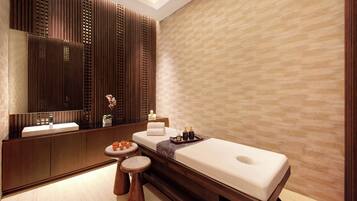 A modern spa room with a wooden panel wall, a large mirror, a massage table, a small round table with candles, and a wooden bench.