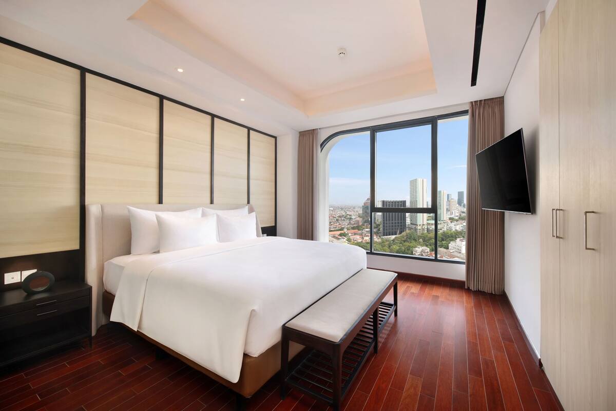 A modern hotel room with a large bed, a bench, a TV, and a city view.