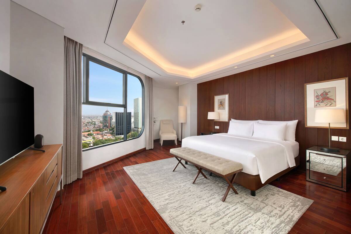 A modern hotel room with a large bed, a TV, a bench, a desk, and a view of the city.