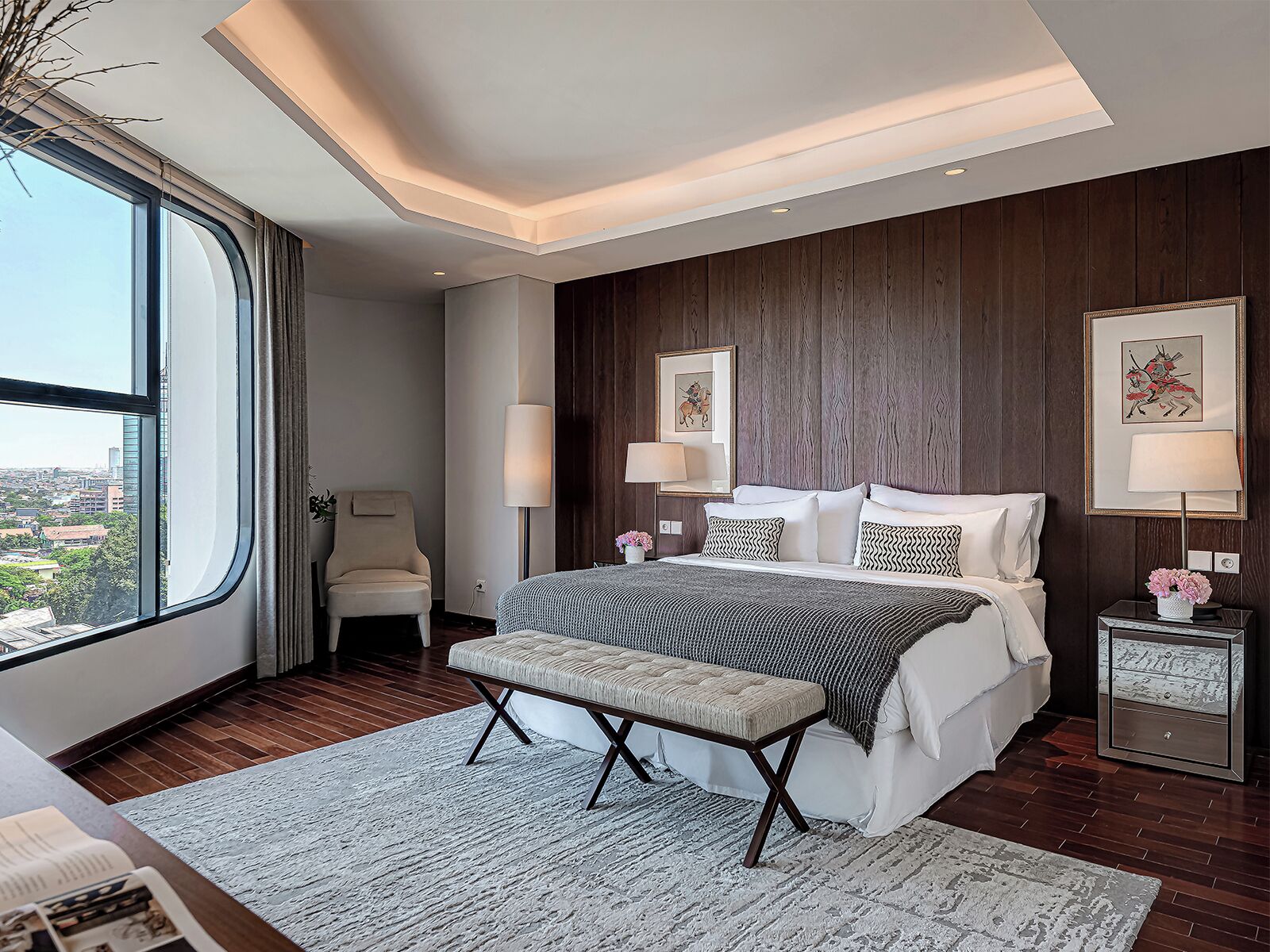 A modern bedroom with a large window, a bench, a bed with patterned pillows, and two wall-mounted lamps.