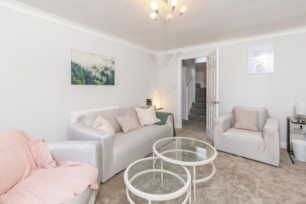 Un salon comprenant un canapé, deux fauteuils, une table basse en verre, un lustre et un escalier menant à une autre pièce.