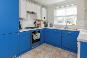 Une cuisine avec des meubles de rangement bleus, une cuisinière à gaz, un four, un évier et une fenêtre avec des stores.