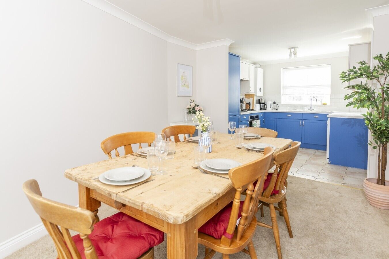 Een eetgedeelte met een houten tafel, gedekt voor zes personen, omringd door houten stoelen met rode kussens. Op de achtergrond is een keuken met blauwe kastjes zichtbaar.