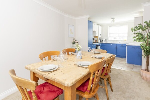 Comedor con mesa de madera dispuesta para seis personas, rodeada de sillas de madera con cojines rojos. Se observa una cocina con armarios azules al fondo.