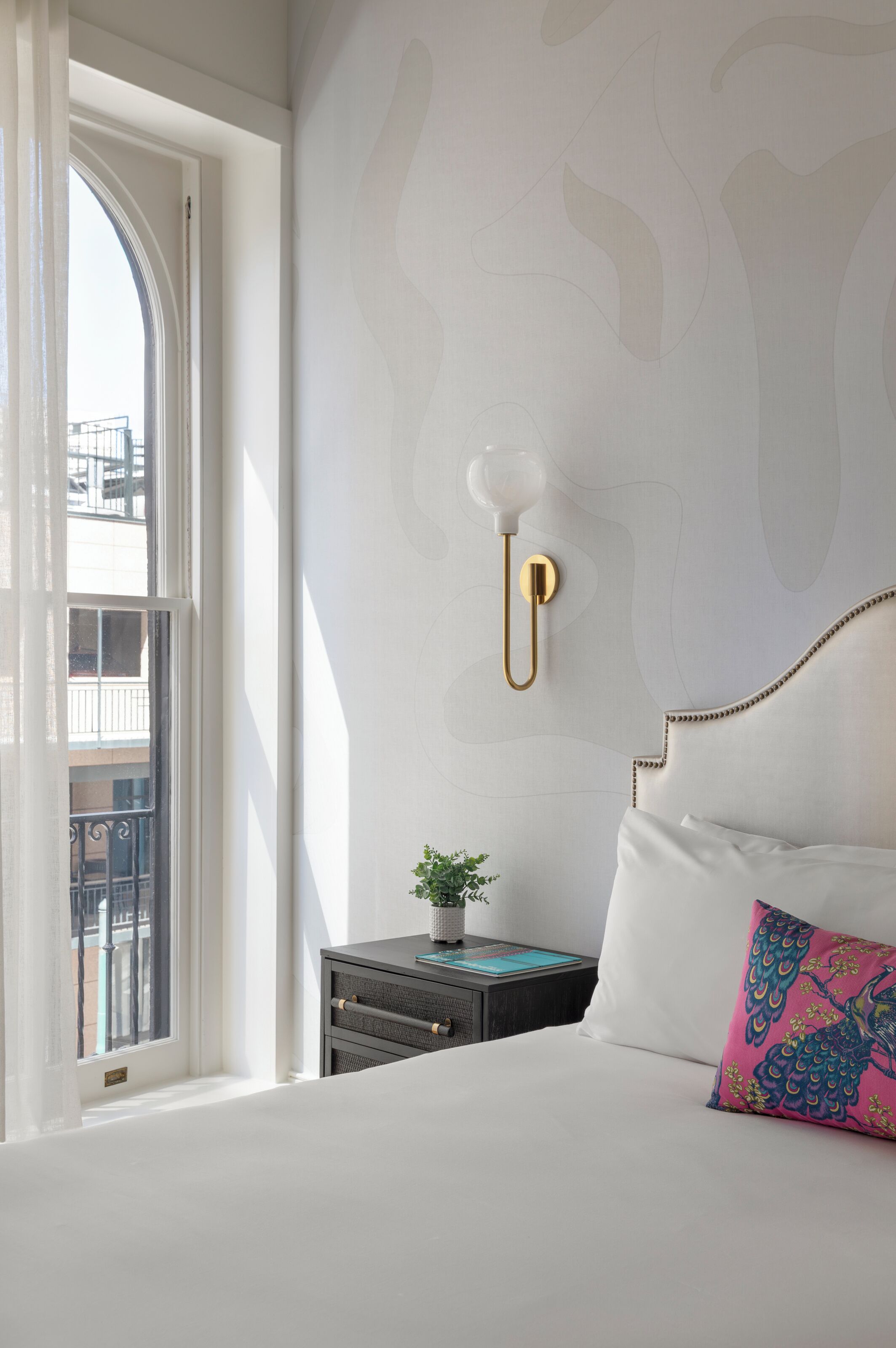 A bedroom with a large window, a bedside table with a plant, a decorative wall light, and a bed with a colorful pillow.