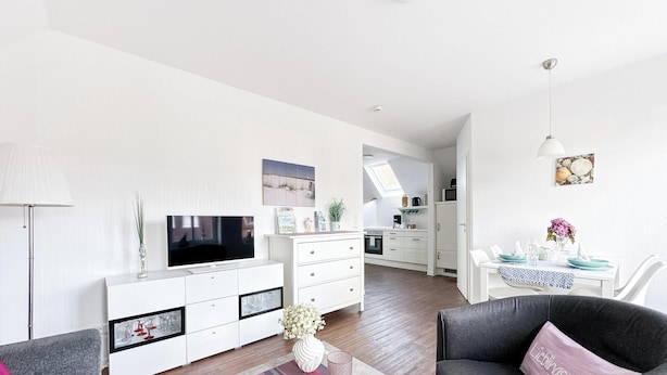 Ein modernes Wohnzimmer mit Flachbildfernseher, weißen Schränken, einem Essbereich mit Tisch und Stühlen sowie einer Küche im Hintergrund.