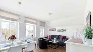 Ein modernes Wohnzimmer mit einer Couch, einem Esstisch, Stühlen und einem Couchtisch. Große Fenster, eine Deckenleuchte und eine Pflanze sind rechts zu sehen.