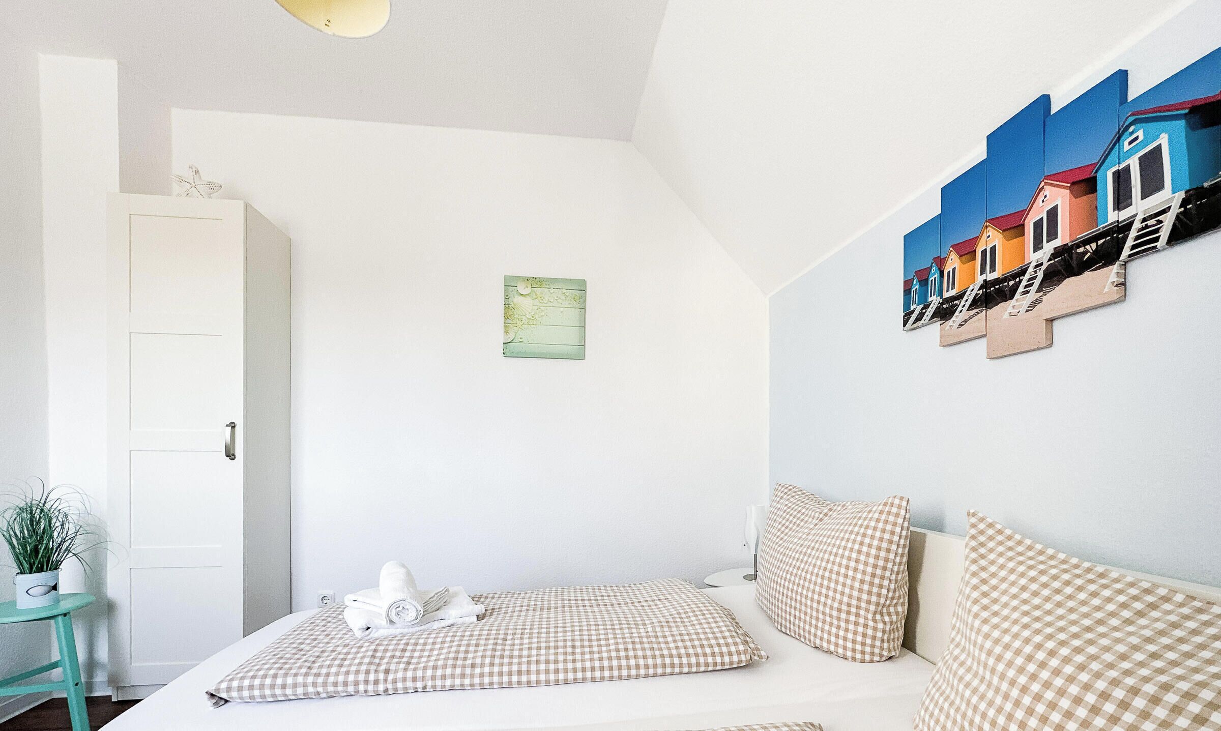 A bedroom with a bed, checkered pillows, a small table with a plant, a white wardrobe, and a wall with colorful artwork.