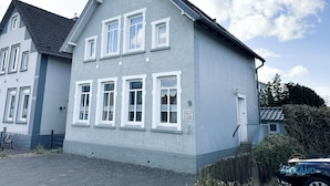 Ein zweigeschossiges Gebäude mit grauer Fassade, weißen Fensterrahmen und einem kleinen Rundfenster im zweiten Stock.