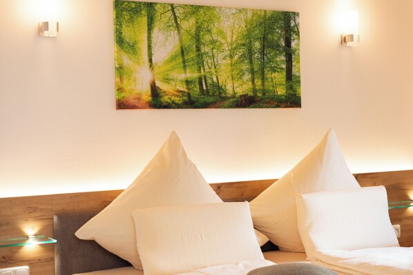 A hotel room with a bed, two white pillows, a wall-mounted painting of a forest, and two wall sconces.