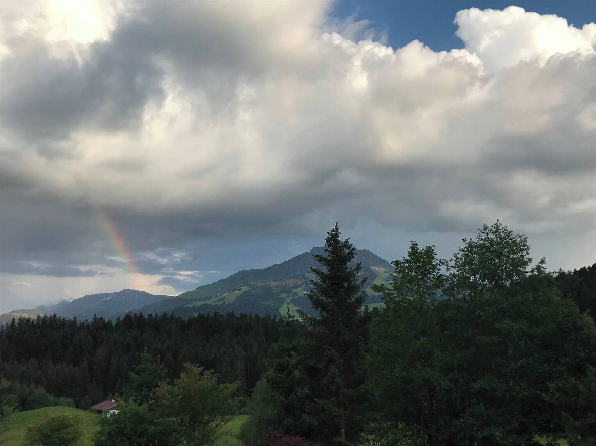 A mountain landscape with a rainbow, dense forest, and a small building.