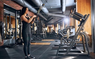 Un gimnasio con equipamiento de ejercicio, una persona usando una máquina de poleas y un banco de pesas.